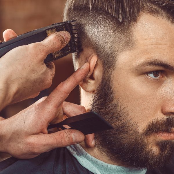 The hands of young barber making haircut of attractive bearded man in barbershop